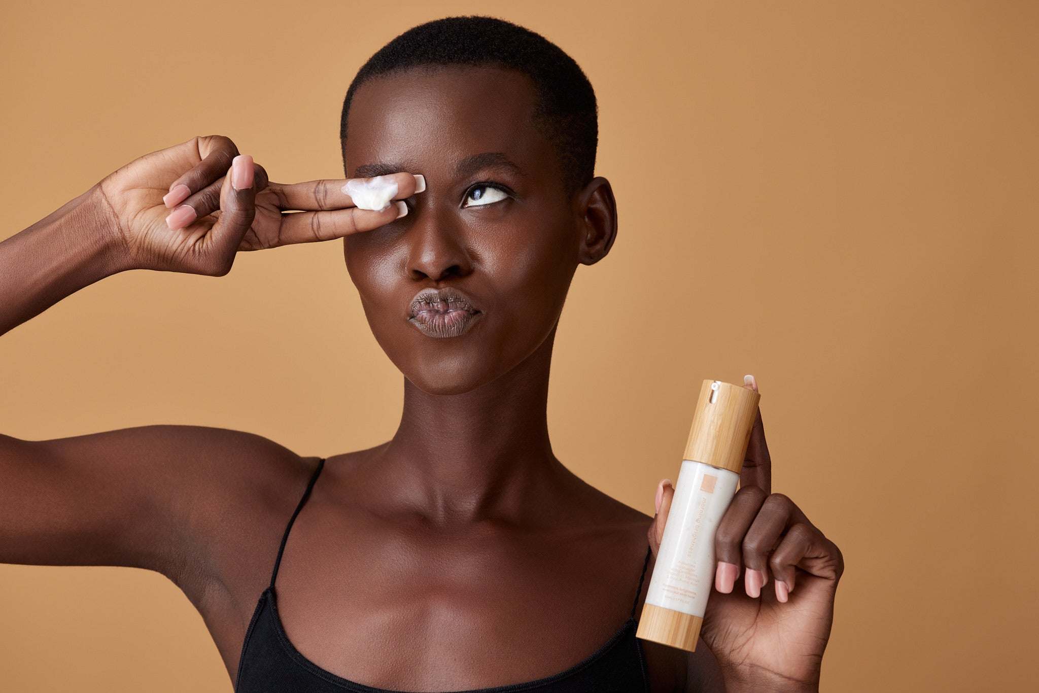 Woman holding the Morning Brightness moisturizer bottle while a small amount of cream is on her finger over her eye.