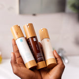 Hands holding three facial moisturizer bottles with bamboo caps, including Morning Brightness, in a bathroom