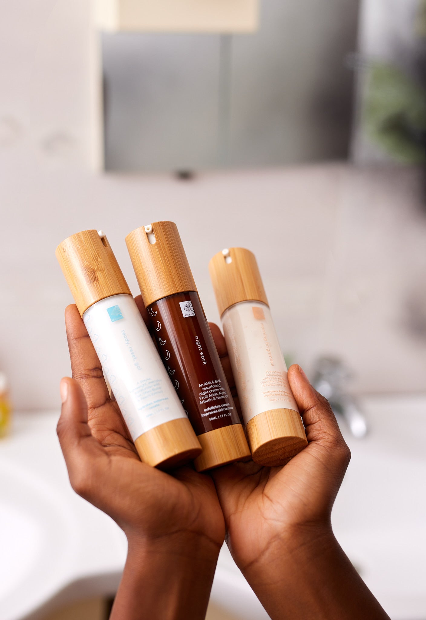 Hands holding three facial moisturizer bottles with bamboo caps, including Morning Brightness, in a bathroom