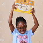 A young girl with braided hair smiles while holding up a box of nokware skincare littles bath soaps against a beige background filled with floating bubbles. She is wearing a light blue graphic t-shirt.