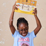 A young girl with braided hair smiles while holding up a box of nokware skincare littles bath soaps against a beige background filled with floating bubbles. She is wearing a light blue graphic t-shirt.
