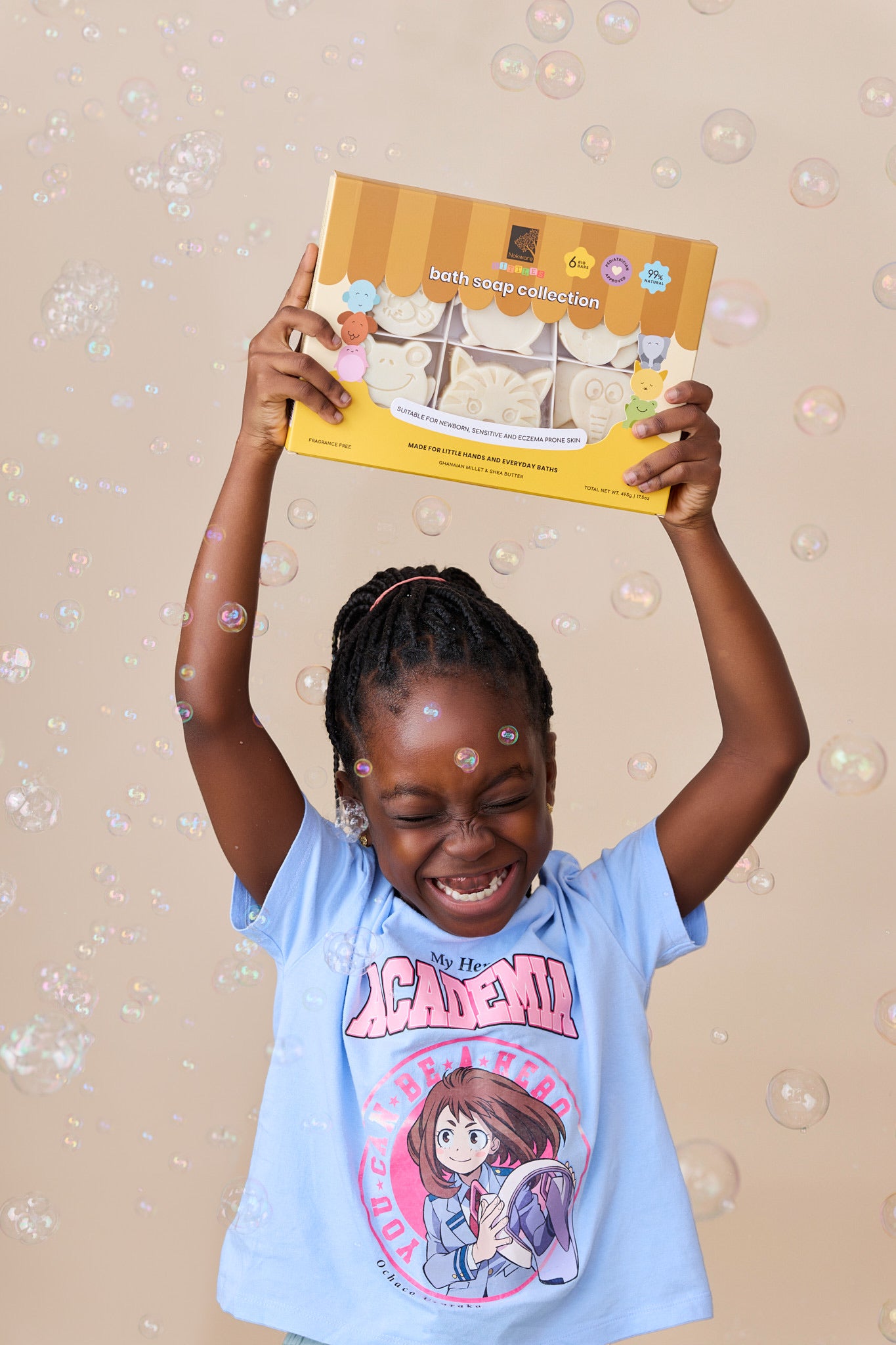 A young girl with braided hair smiles while holding up a box of nokware skincare littles bath soaps against a beige background filled with floating bubbles. She is wearing a light blue graphic t-shirt.